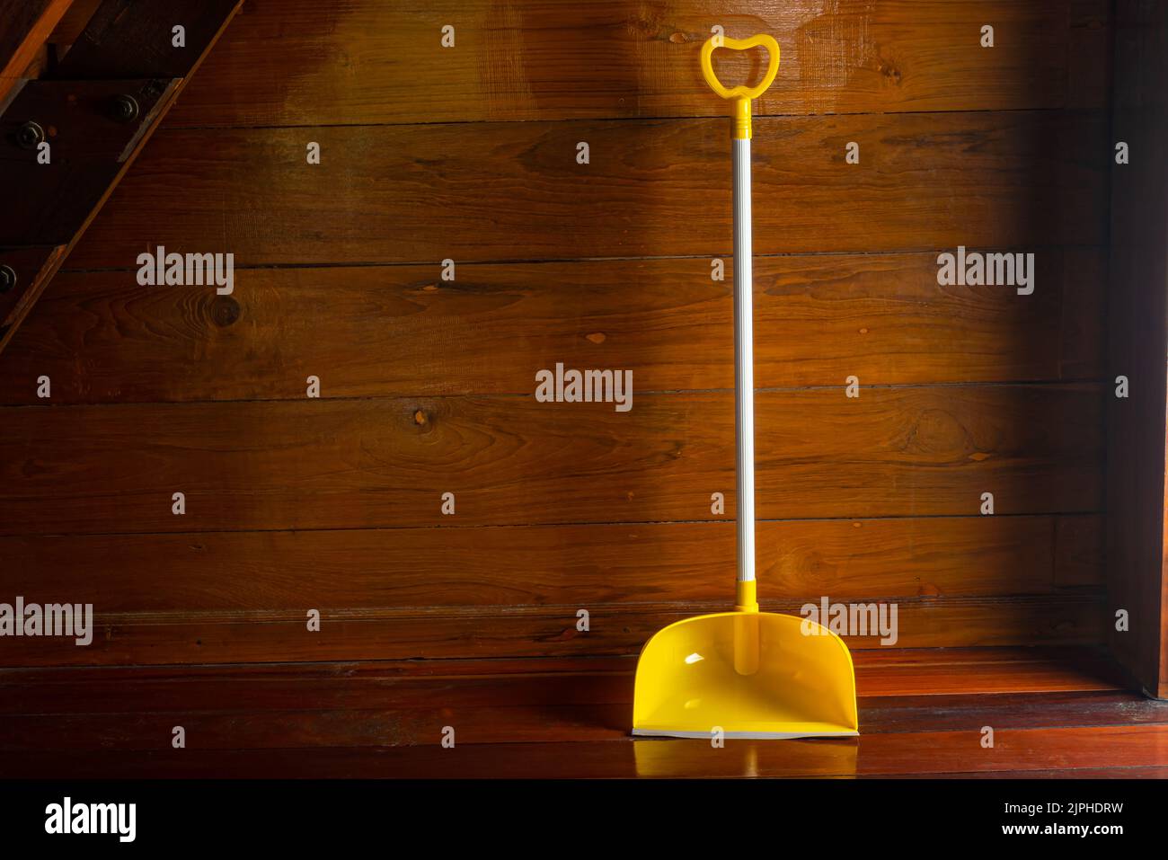Long handle plastic yellow dustpan leaning against wooden wall on a wooden floor in a room. Stock Photo