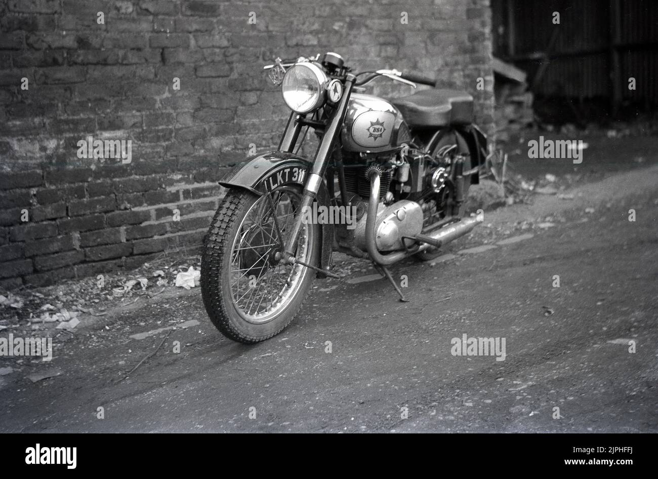 1950s, historical, a British made BSA motorcycle of the era parked on a dirt path outside an old brick warehouse, England, UK. Metal RAC badge on front fork. Founded in 1861 as the Birmingham Small Arms Company Ltd, for the production of firearms, a motorcycle division was established in 1903, and the first motorcycle released in 1910. In the 1950s, BSA Motorcycles was the largest motorcycle maker in the world. Stock Photo