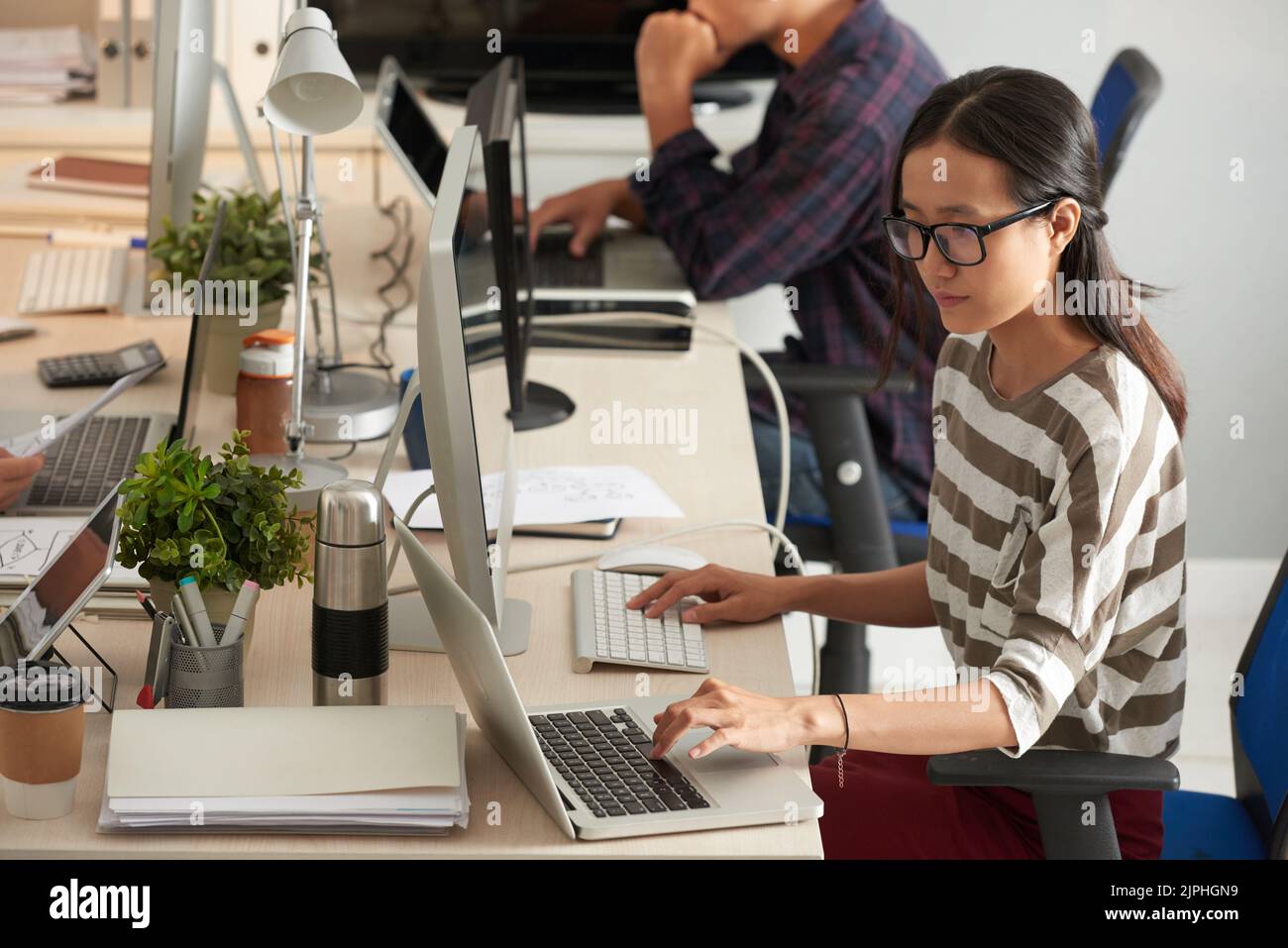 Working process at modern open plan office: pretty young manager in casualwear analyzing statistic data while using modern computer and laptop, profile view Stock Photo