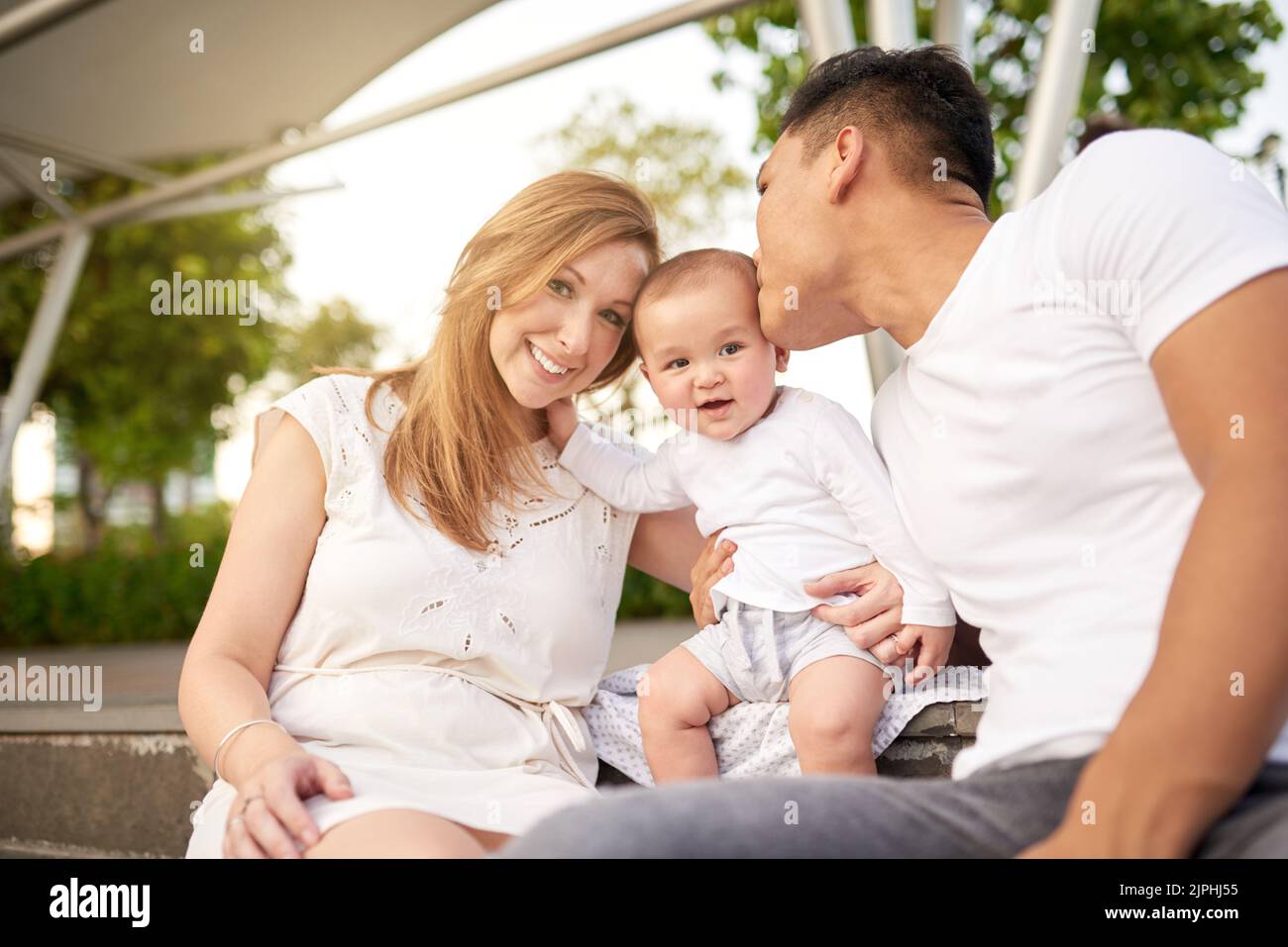 Group portrait of loving mixed-race parents sitting at green public park and cuddling their cute little son, attractive Caucasian woman looking at camera with wide smile Stock Photo