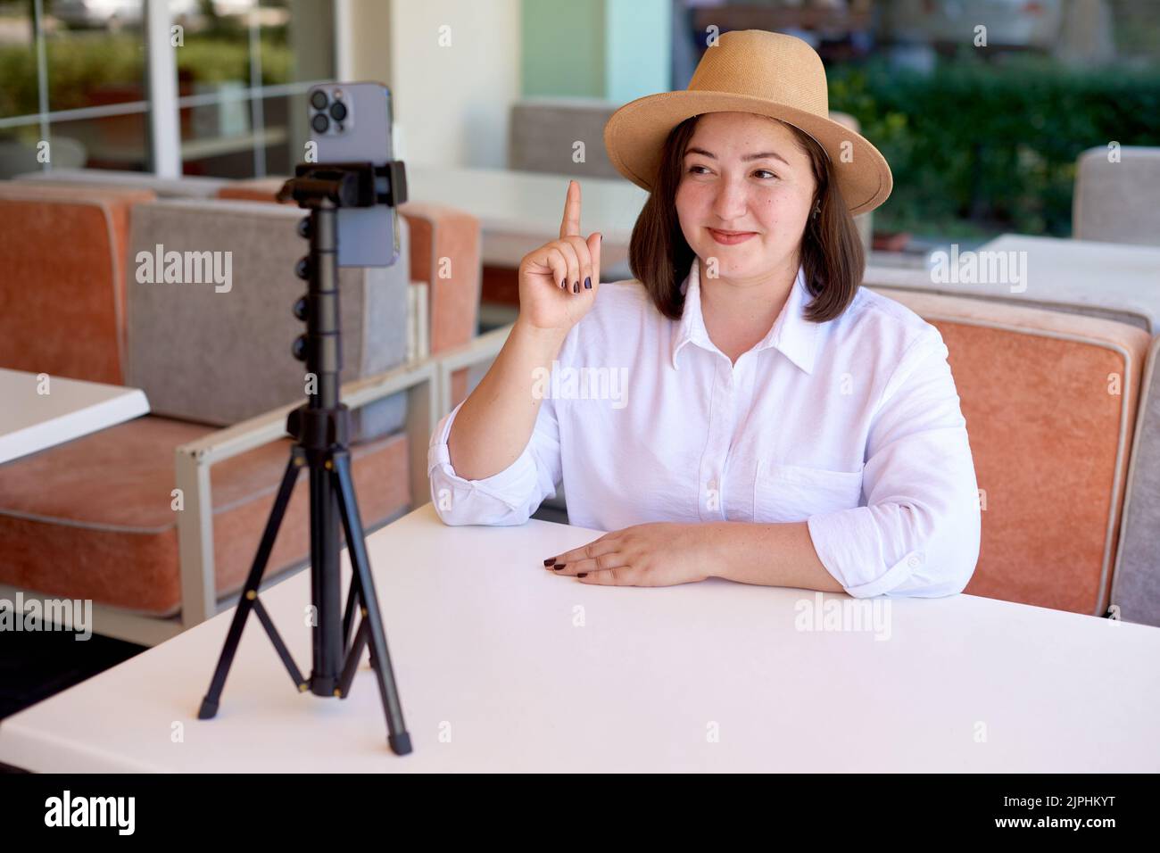woman bends her fingers and counts out loud number one on camera phone. woman online with mobile phone on tripod streaming in summer terrace cafe. sha Stock Photo