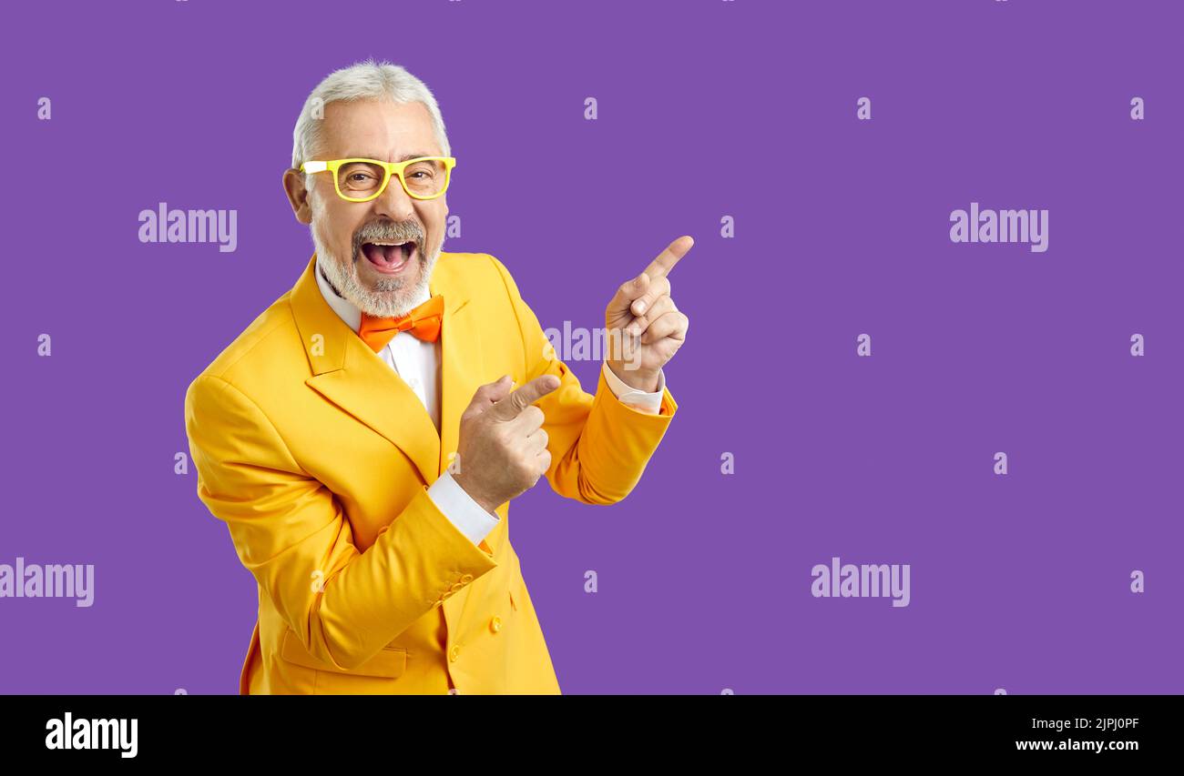 Cheerful extravagant and funny senior man pointing fingers at copy space on purple background. Stock Photo