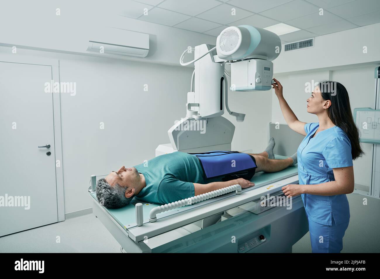 Male patient lying on bed while female nurse adjusting modern X-ray machine for scanning his leg or knee for injuries and fractures Stock Photo