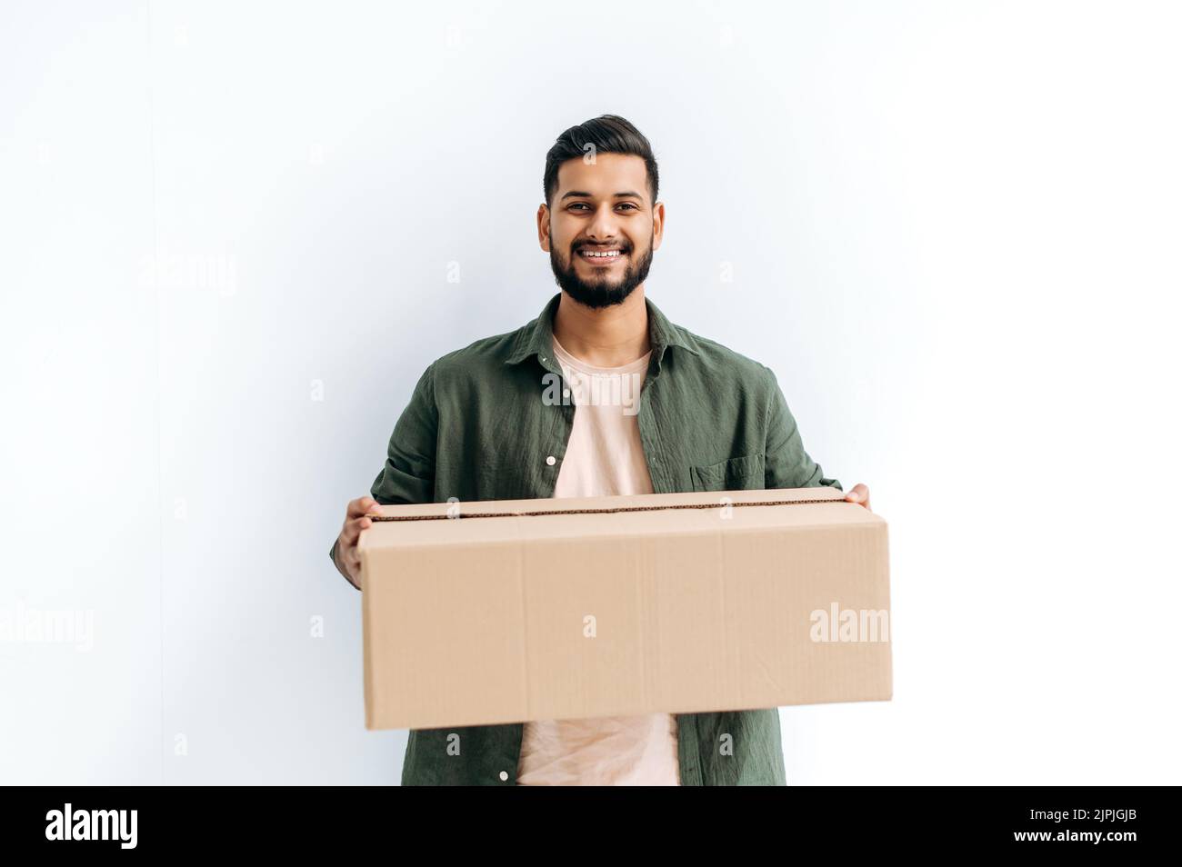 Positive attractive candid arabian or indian young delivery man in casual clothes, holding a cardboard box, standing on isolated white background, looks at camera, smiles friendly. Delivery concept Stock Photo