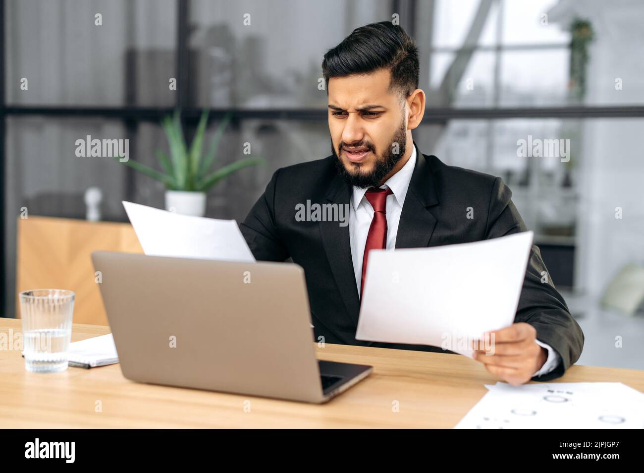 Disappointment stressed indian or arabian business man, sit at work desk with laptop in modern office, works with documents, upset with financial report, analyzes the result of work, feels frustrated Stock Photo