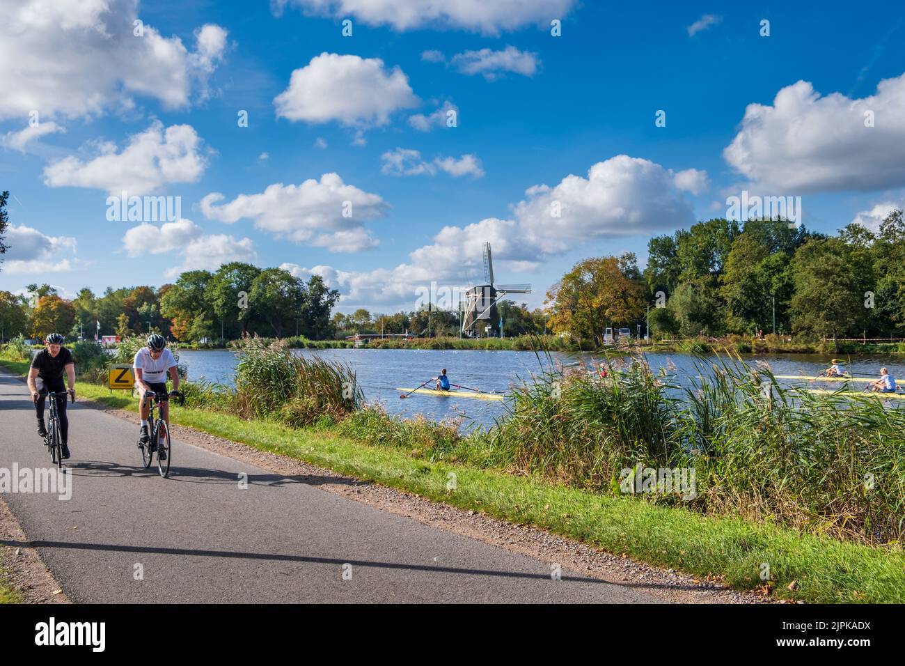 Two Cyclist along the Amstel River wiyh Rieker Windmill in background,  Amsterdam, Netherlands Stock Photo