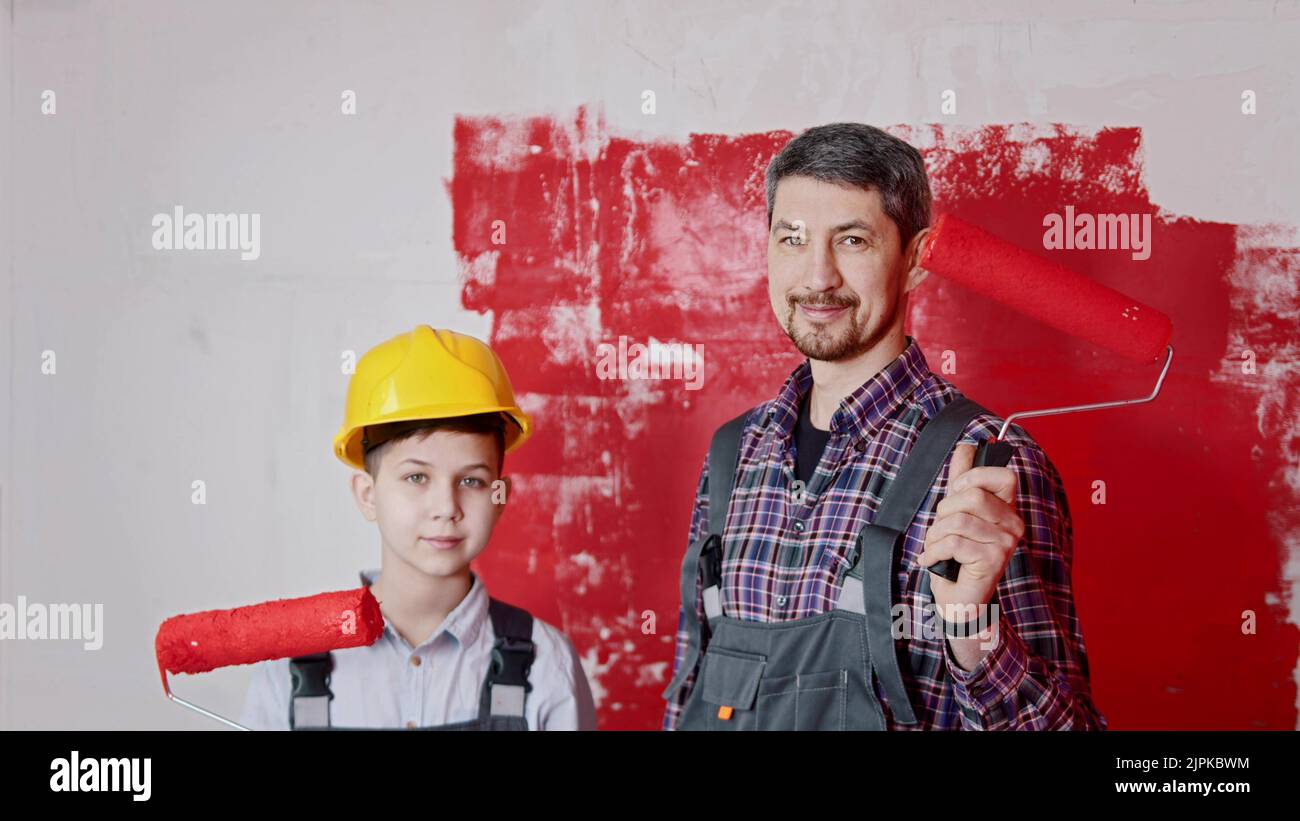 A little boy and his smiling father standing in the draft room and holding rollers covered in red painting Stock Photo