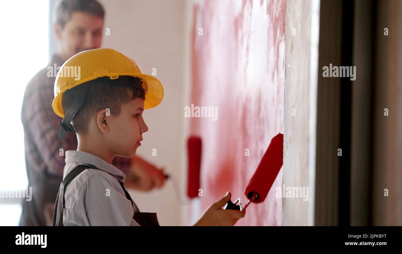 A little boy and his smiling father painting walls in red color - a boy wearing helmet Stock Photo