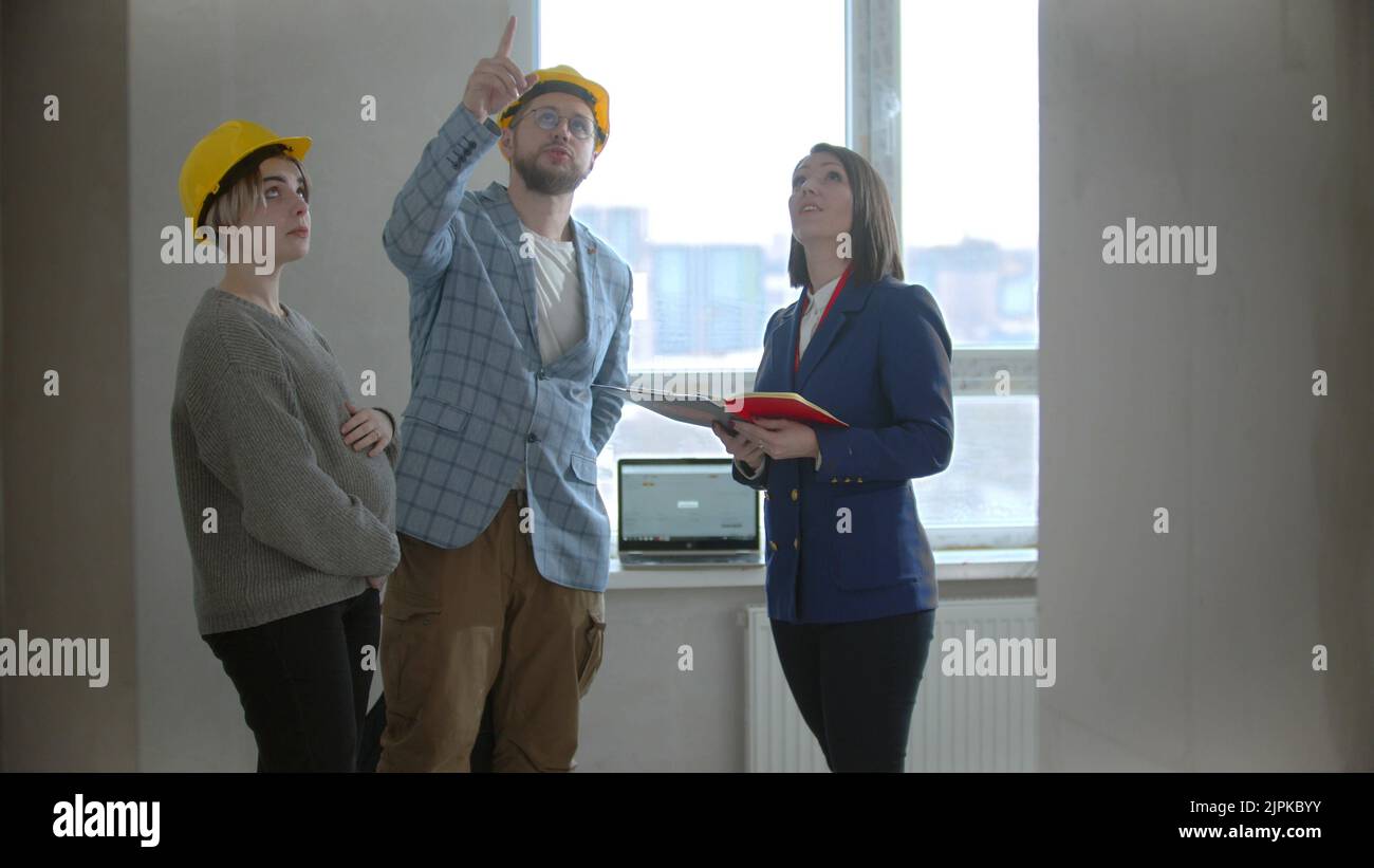 A pregnant woman with her husband talking with a real estate agent in apartment in a new building - man asking questions and pointing at the ceiling Stock Photo