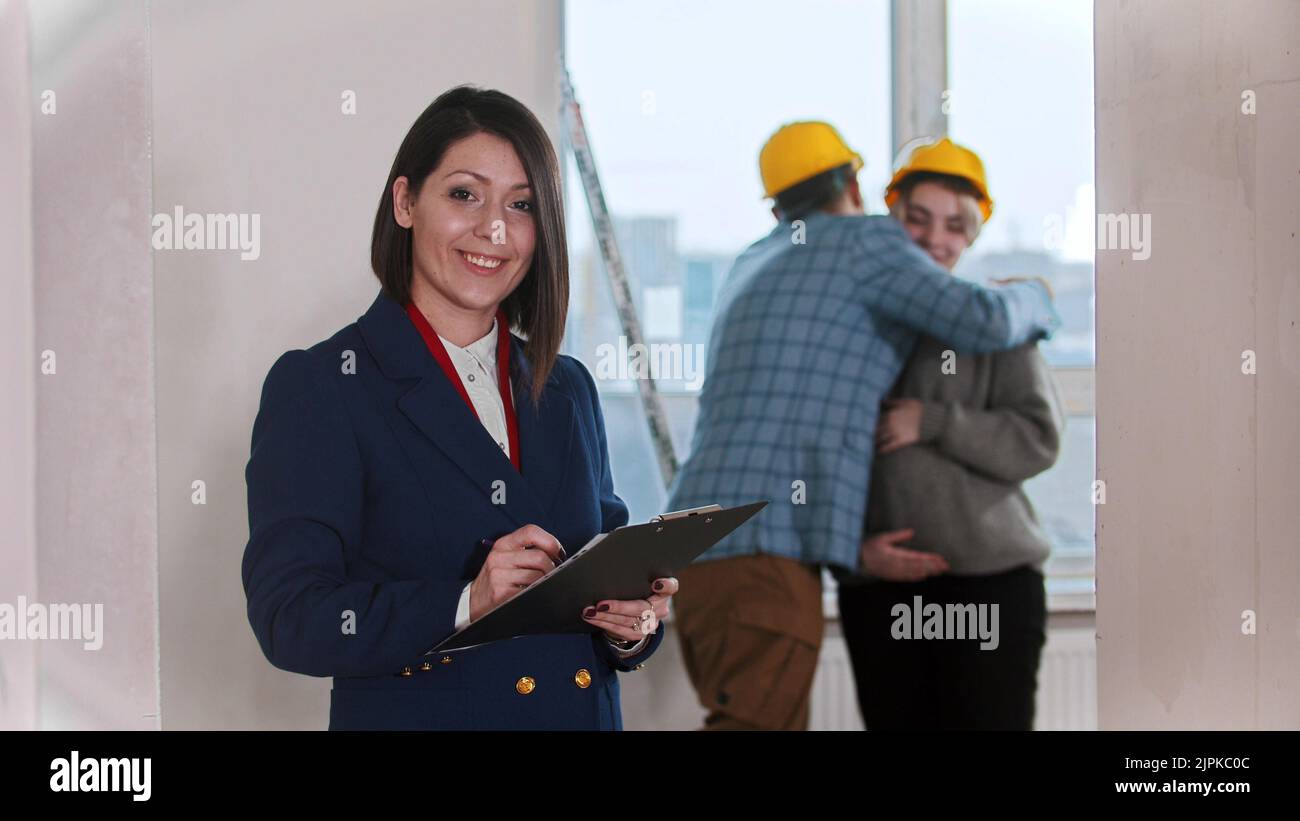 A smiling real estate agent standing in draft apartment - young married couple hugging each other on the background Stock Photo