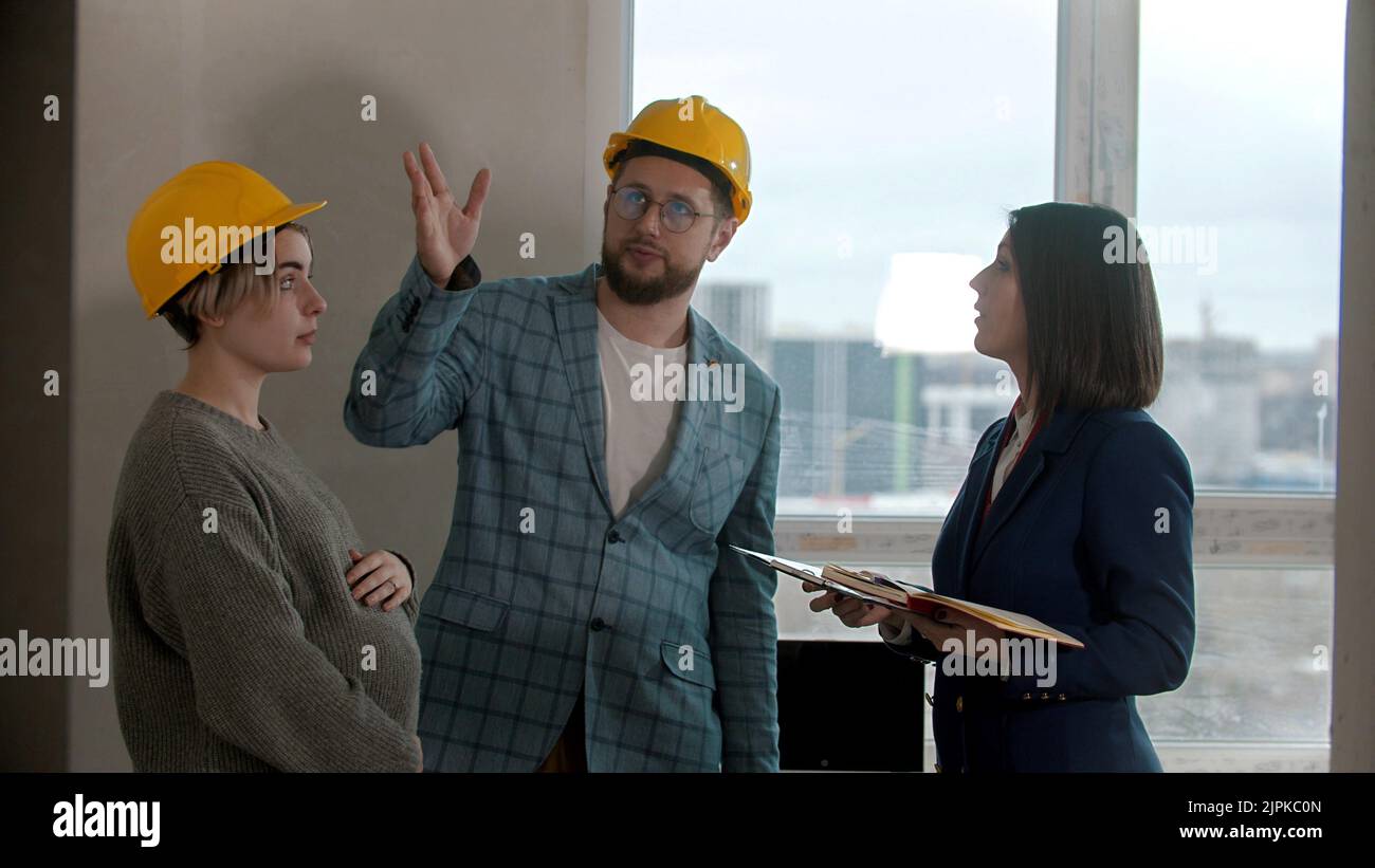 A pregnant woman with her husband talking with a real estate agent in apartment in a new building - man asking questions and pointing at the walls Stock Photo