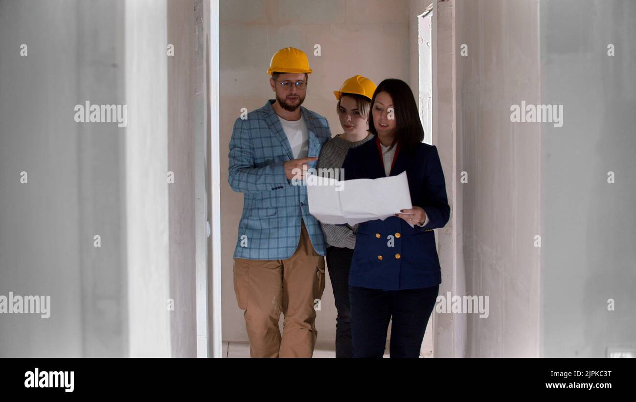 A real estate agent showing a new draft apartment to a young married couple in helmets - looking at the paper Stock Photo