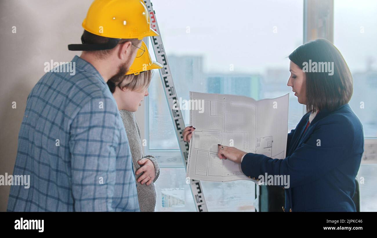 A real estate agent showing a layout to a young married couple in yellow helmets Stock Photo