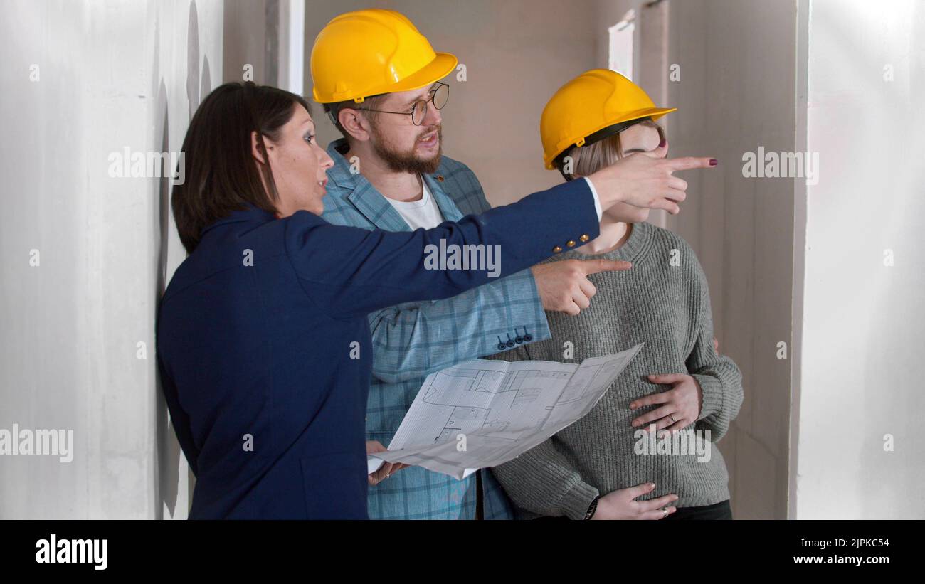 A real estate agent showing a new draft apartment to a young successful married couple holding a layout on the paper Stock Photo