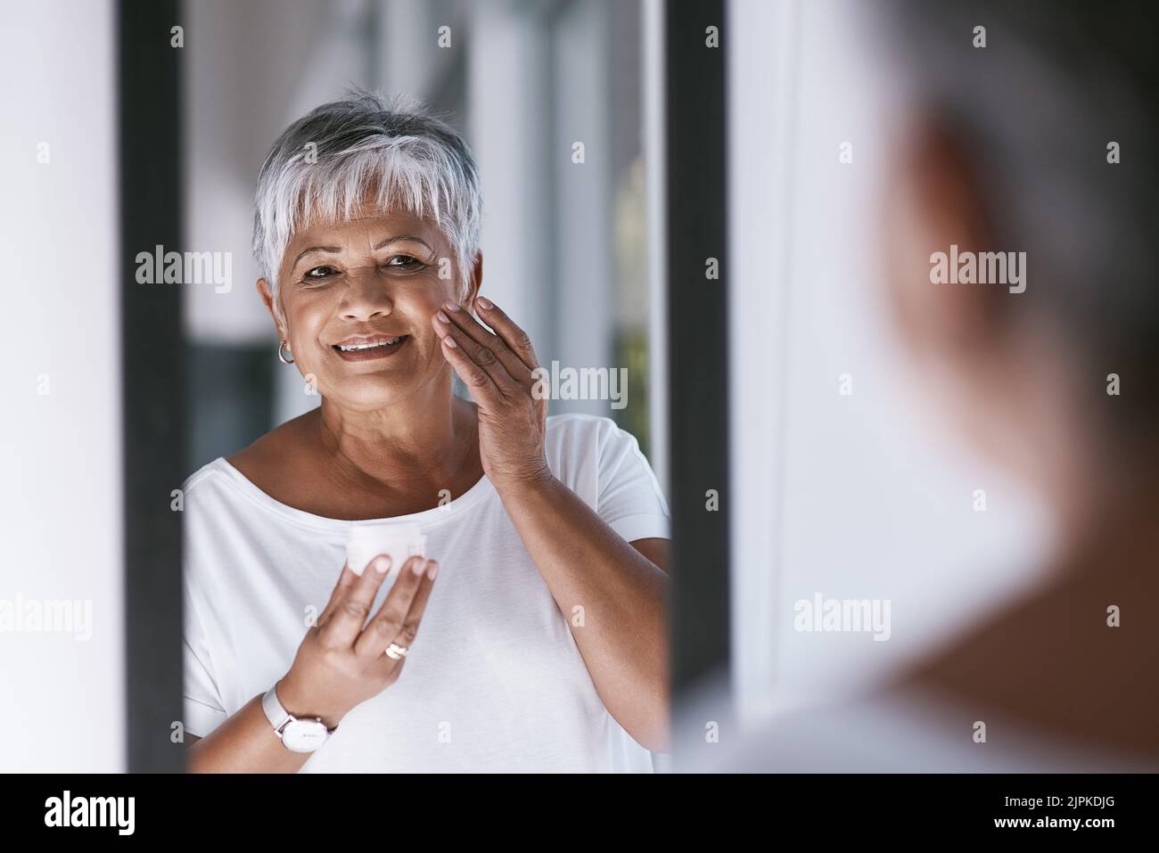 I feel younger. a cheerful mature woman applying skin cream on her face while looking into a mirror at home. Stock Photo
