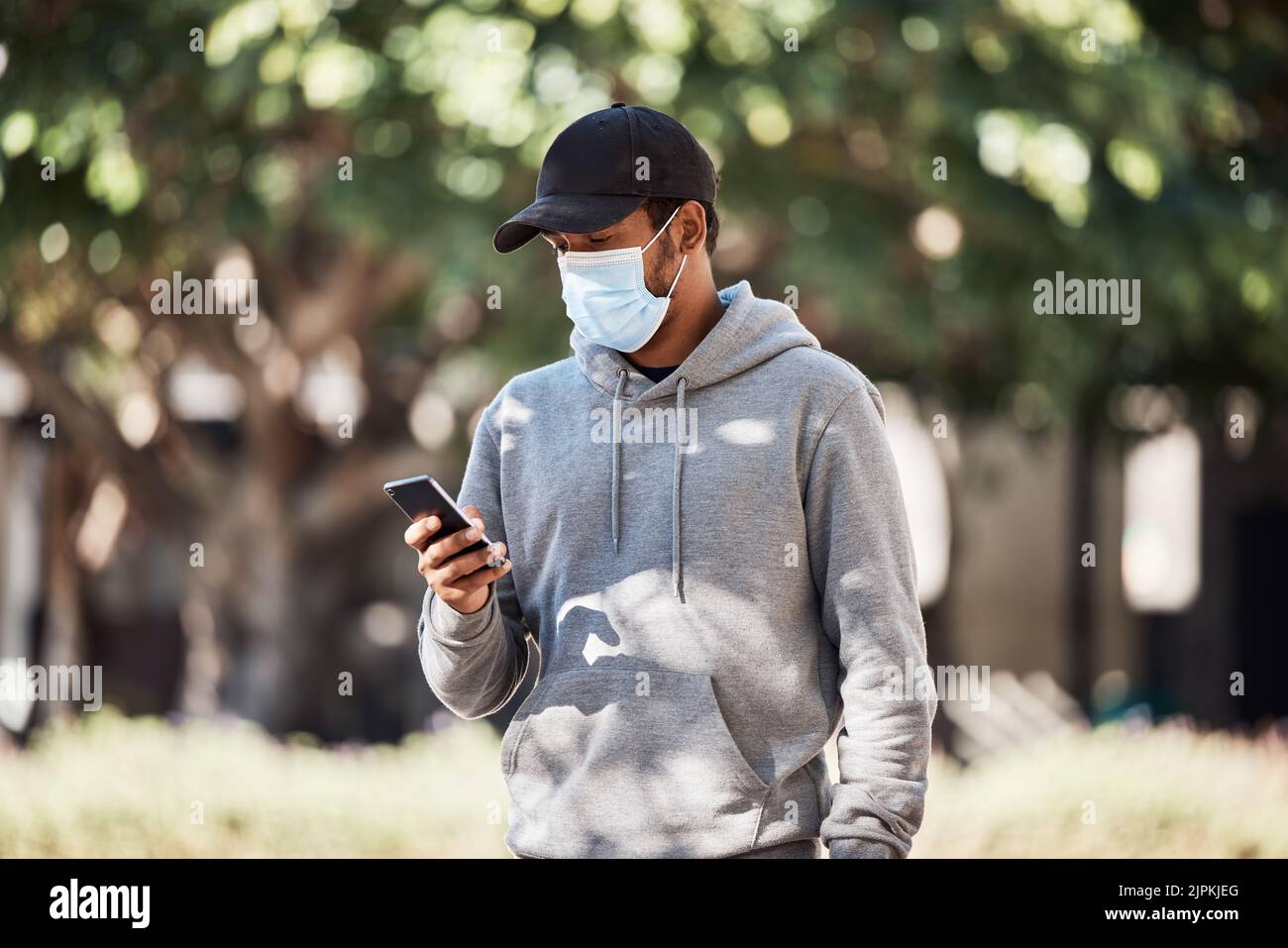 Man looking at phone in nature park, wearing face mask to prevent risk of covid and reading a text message online. Guy standing in garden outside Stock Photo