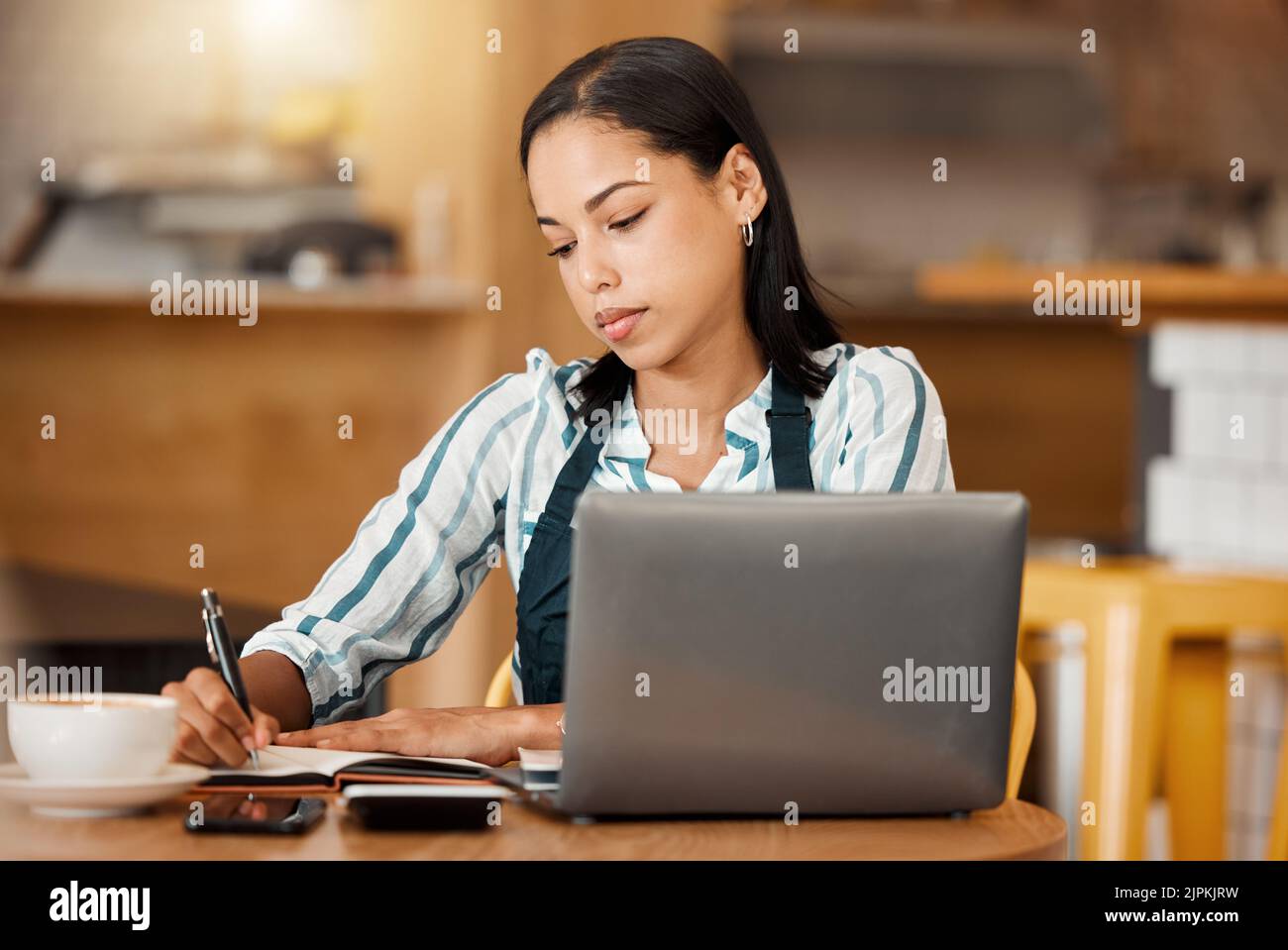 Cafe owner, coffee shop manager or small business entrepreneur writing notes and working on a laptop in her startup. Young female looking serious Stock Photo