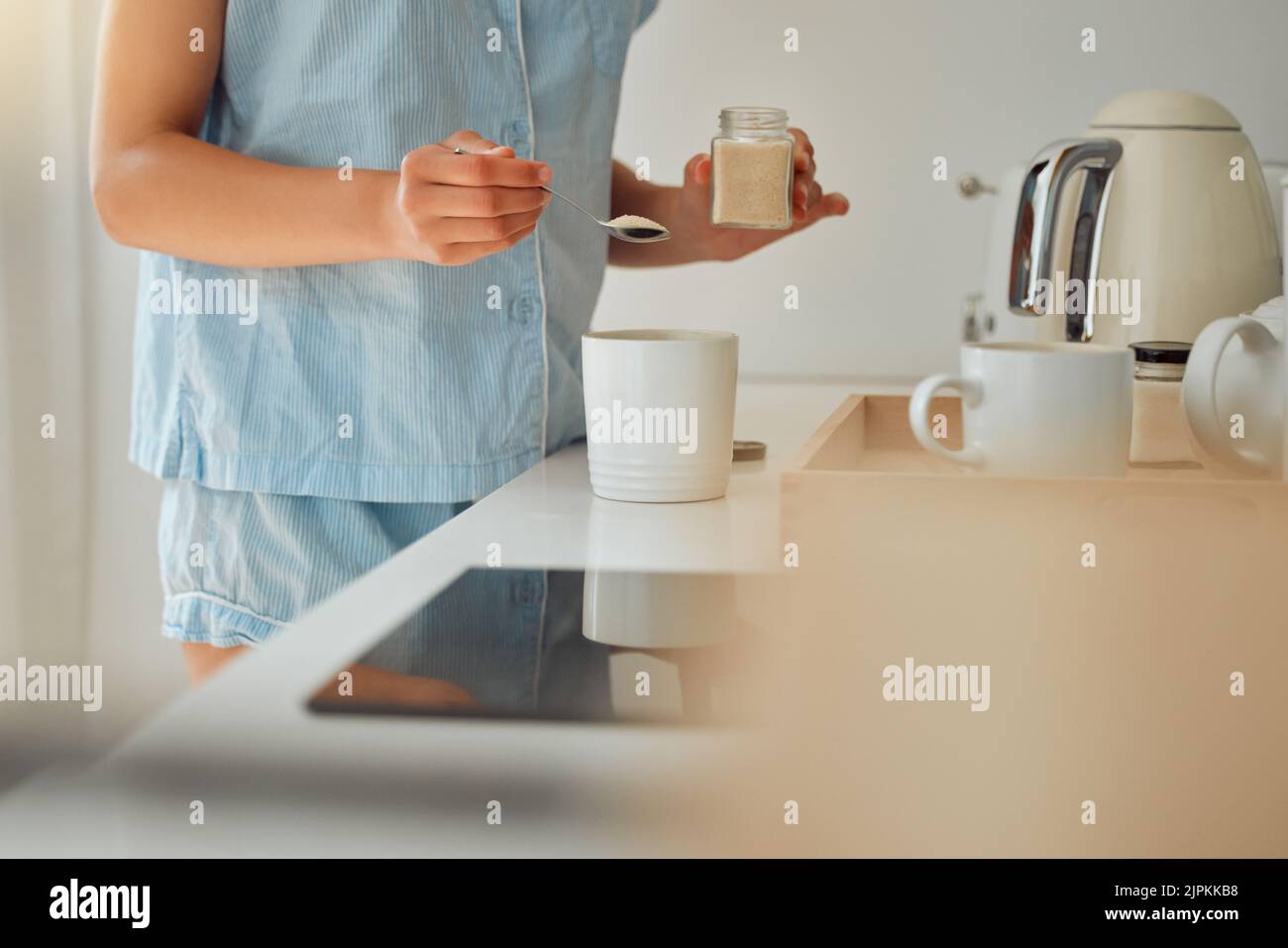Casual woman adding sugar while preparing a cup of coffee, tea or hot chocolate in the kitchen during the morning. Female hands holding teaspoon for a Stock Photo