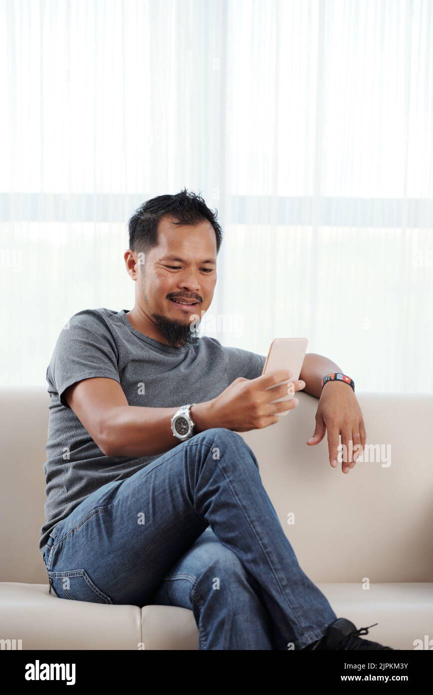 Handsome Vietnamese man with smartphone resting on sofa Stock Photo