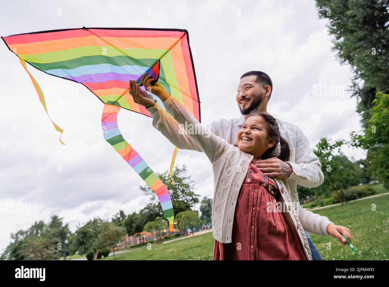 Positive asian dad hugging daughter with flying kite in summer park ...