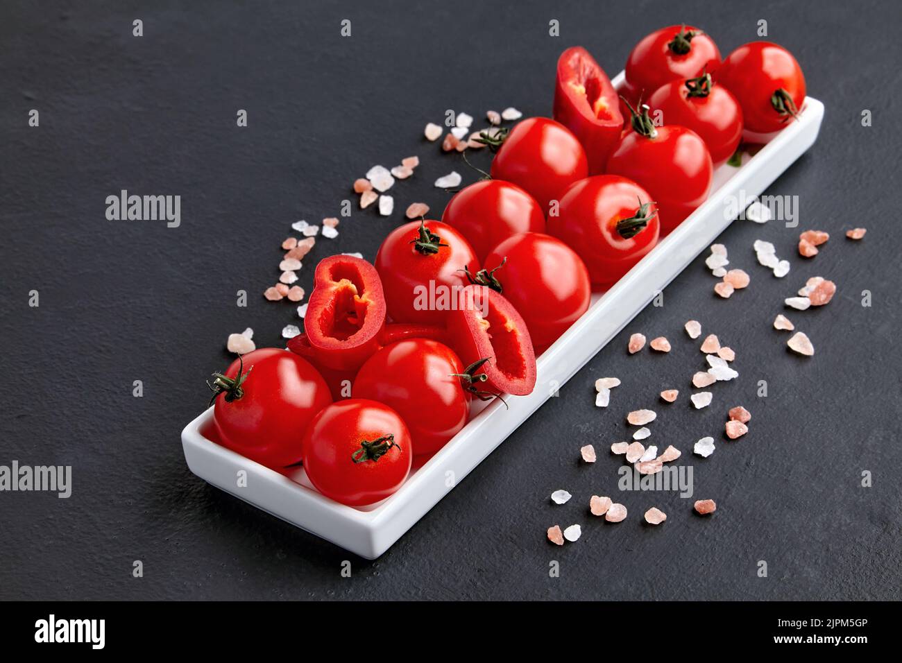 An elongated dish of tomatoes, paprika and coarse pink Tibetan salt on a black concrete table. High angle with copy space Stock Photo