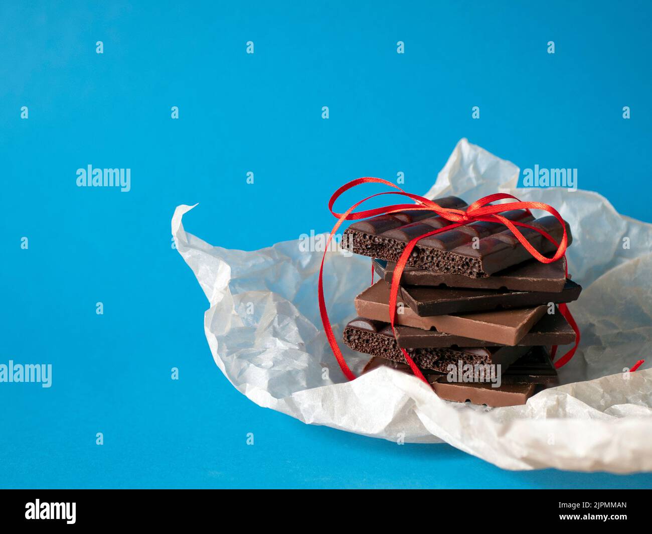 Chocolate stack wrapped up with a red ribbon on blue background, copy space. Concept of sweet present, pleasant gift Stock Photo