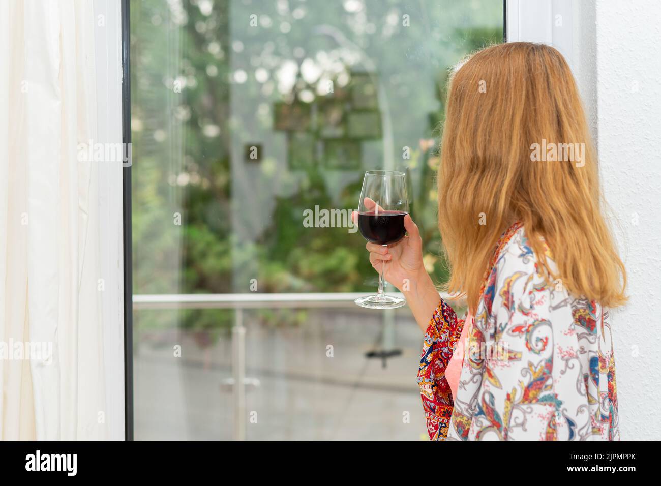 A woman in a colorful dressing gown stands at the window with a glass of drink Stock Photo
