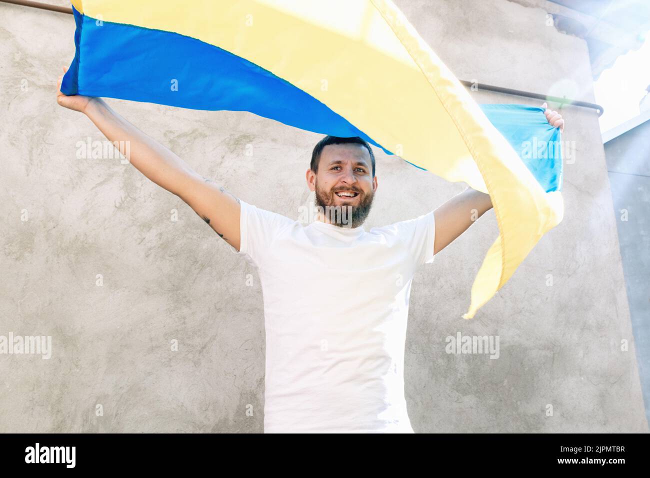 Soft selective focus on Man holding the National Blue and Yellow Flag of Ukraine above his head and smiling. Guy in white T-shirt and jeans. Sunny day. Constitution and Independence Day of Ukraine. Stock Photo