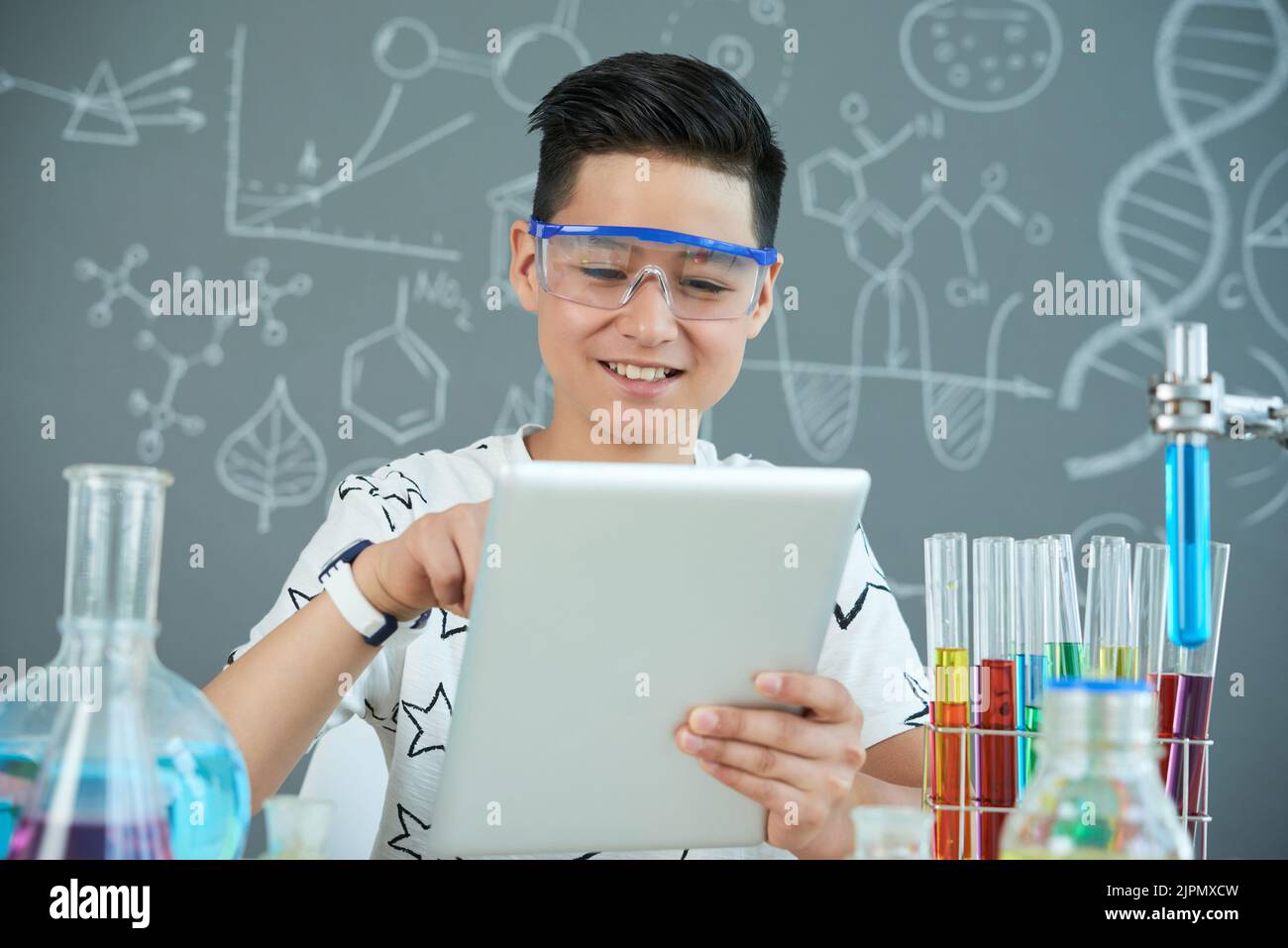 Cheerful Asian pupil wearing protective goggles using digital tablet while finishing school project at modern chemistry class, portrait shot Stock Photo