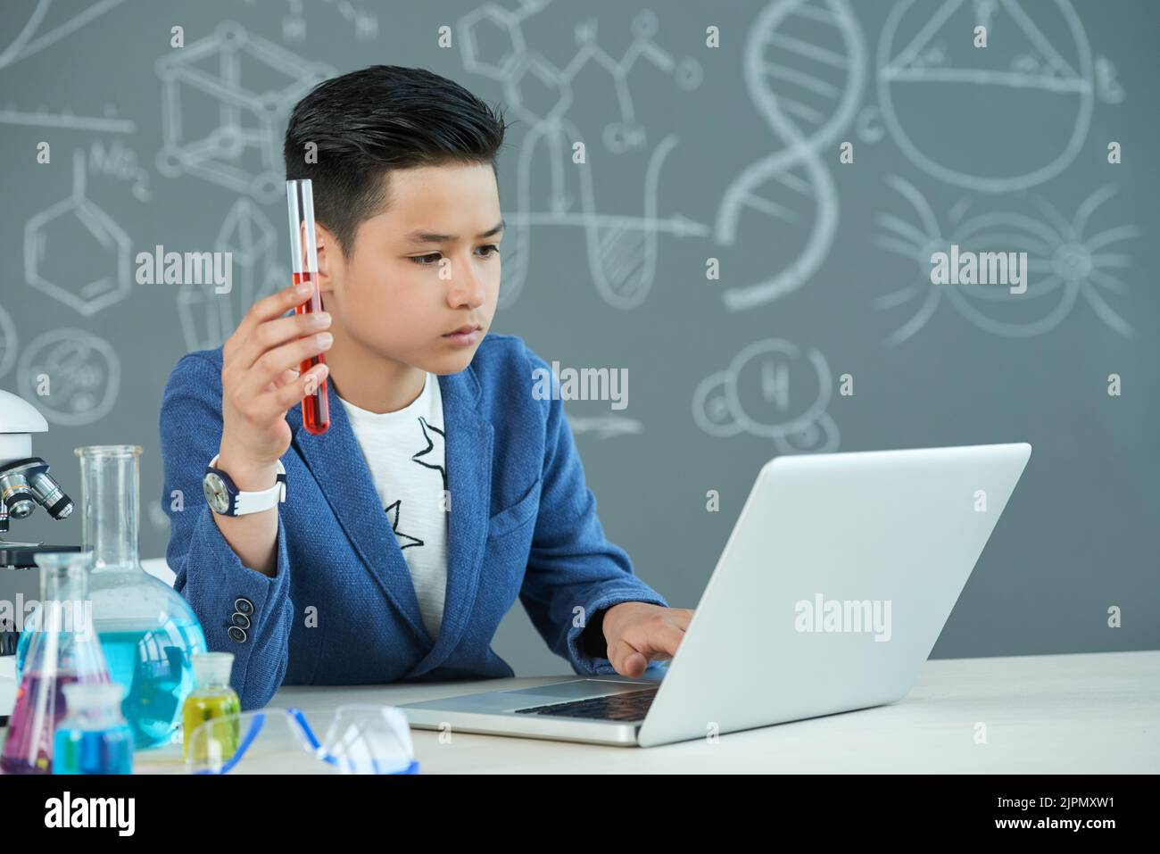 Confident Asian student sitting in front of modern laptop and finishing school research project at chemistry classroom, he holding test tube in hand Stock Photo
