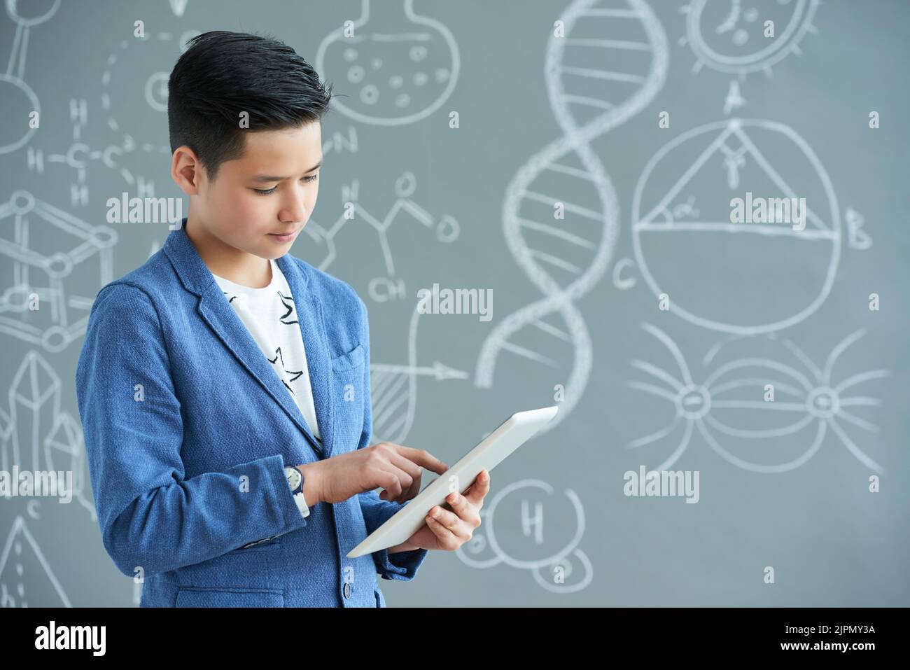 Concentrated Asian student wearing jacket and T-shirt finishing school project with help of modern digital tablet while standing against blackboard, portrait shot Stock Photo