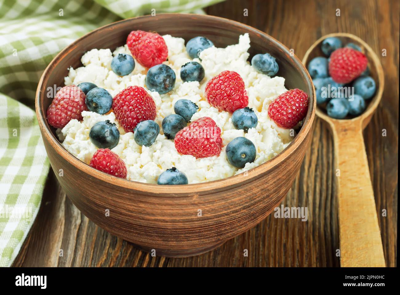 cottage cheese with raspberries and blueberries in brown clay bowl and wooden spoon on brown wooden background. Dairy products, healthy food. Stock Photo