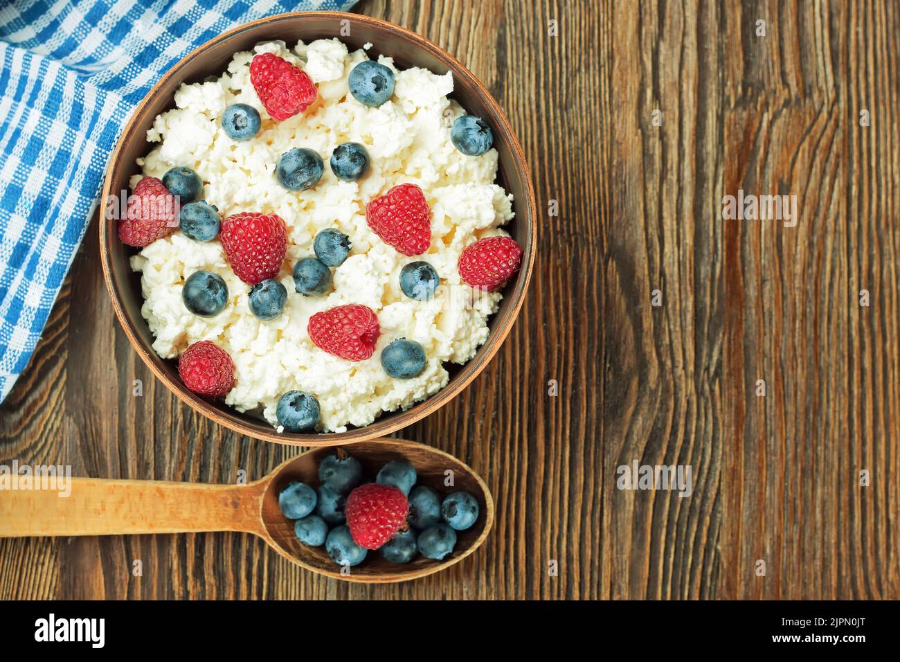 cottage cheese with raspberries and blueberries in brown clay bowl and wooden spoon on brown wooden background. Dairy products, healthy food. Top view Stock Photo