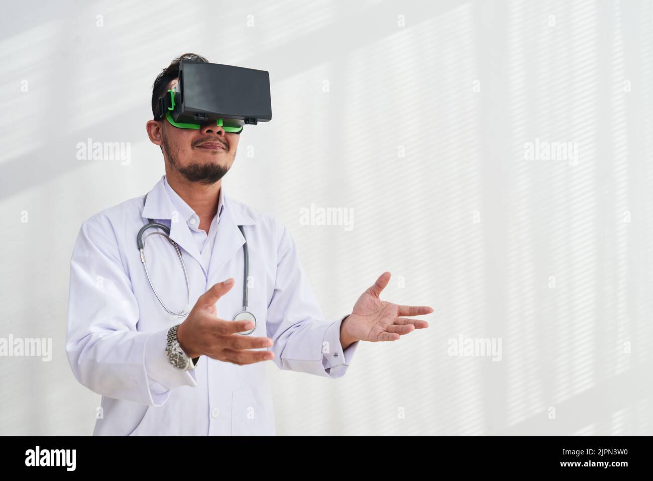 Bearded physician wearing white coat using VR headset while working in modern office, white background Stock Photo