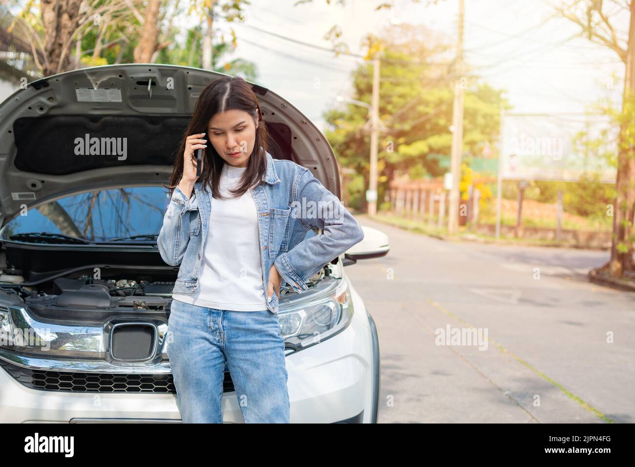 Angry Asian woman and using mobile phone calling for assistance after a car breakdown on street. Concept of vehicle engine problem or accident and eme Stock Photo