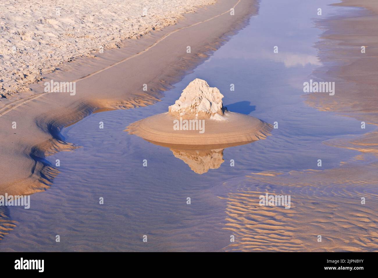 Sand castle on shallow sea water with reflections by morning sunlight. Abstract, surreal beach background. Magic sunrise lights on sand and the water. Stock Photo