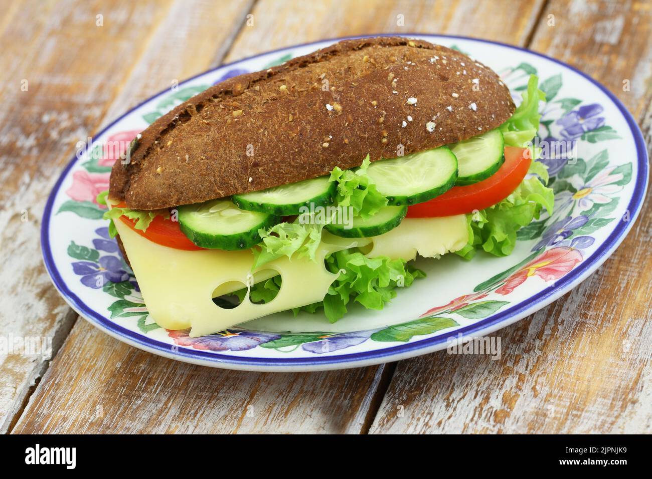 Whole grain roll with Swiss cheese, lettuce, tomato and cucumber on colorful vintage plate on wooden surface Stock Photo