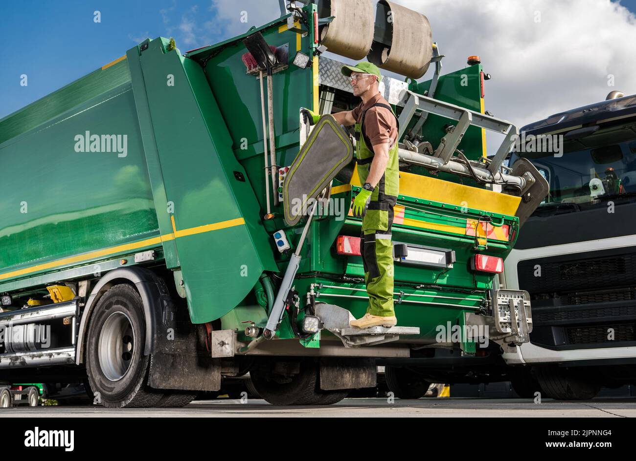 Caucasian Modern Garbage Truck Worker Riding on a Rear Side of the Vehicle. Waste Management Industry Job. Sorting and Recycling Theme. Stock Photo