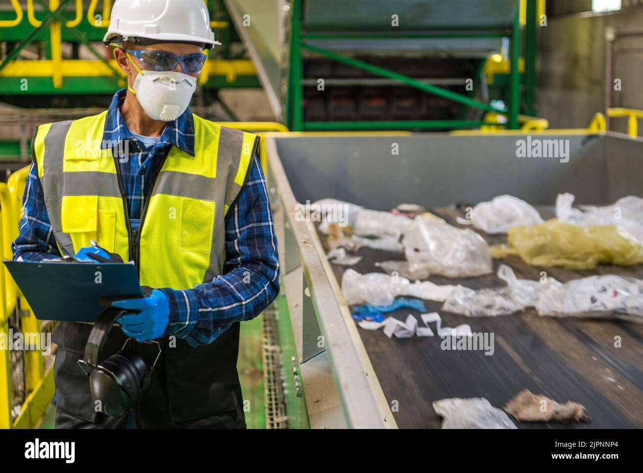 Waste Management Sorting Facility Caucasian Worker Preparing Documentation. Wearing Air Pollution Mask, Eyes Safety Glasses and a Hard Hat. Stock Photo