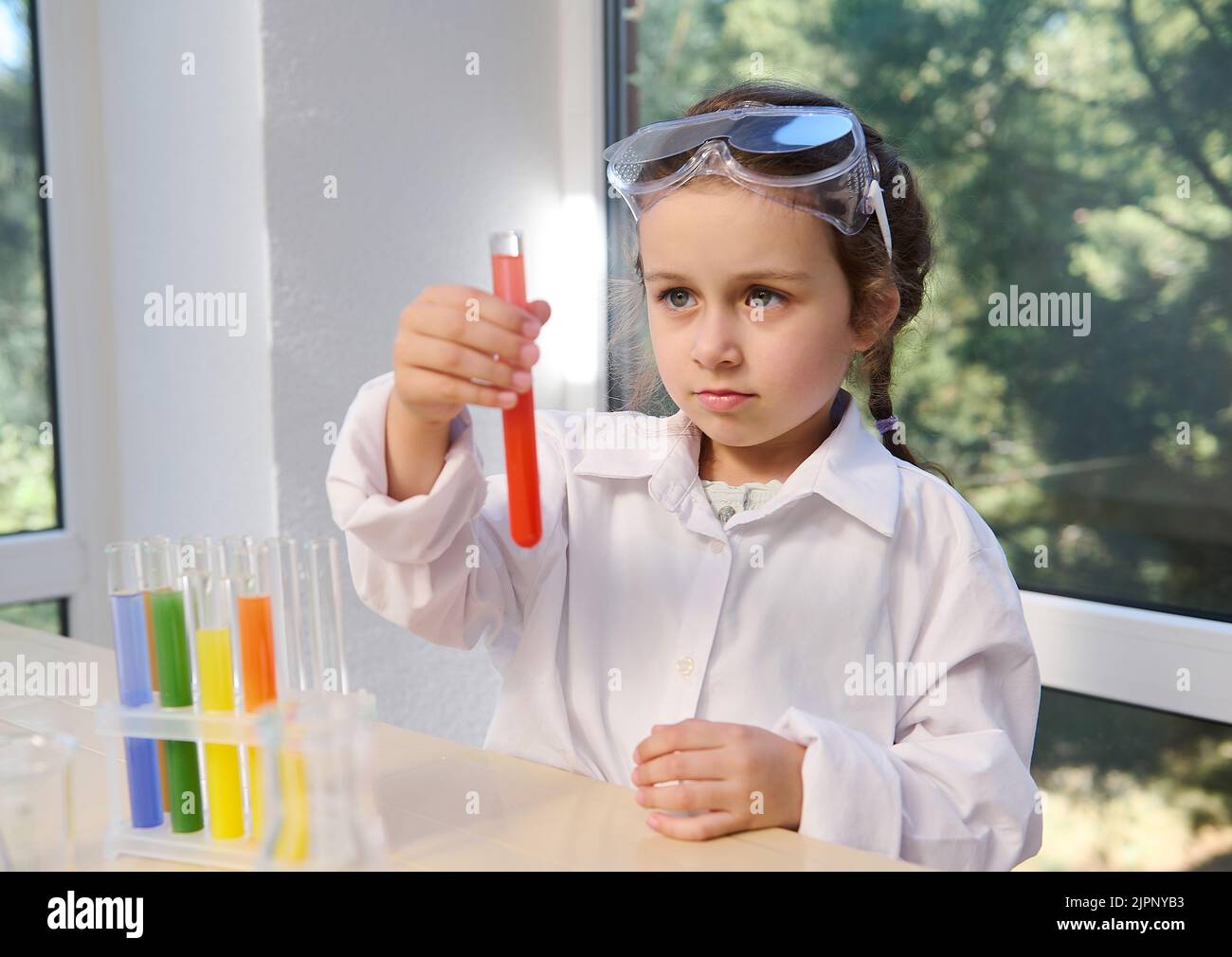 Preschool girl in safety goggles and lab coat watching a chemical