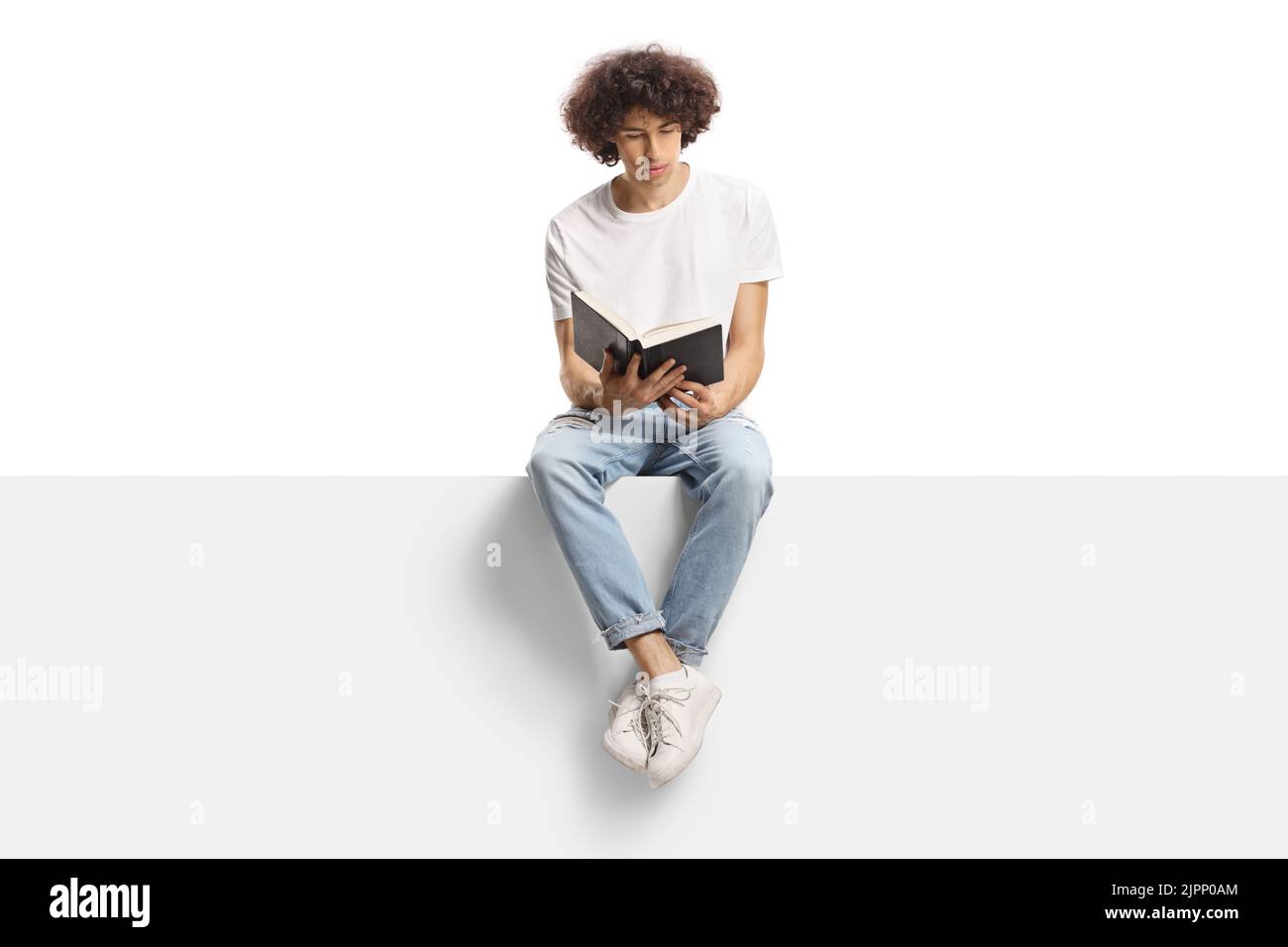 Young man with curly hair sitting on a panel and reading a book isolated on white background Stock Photo