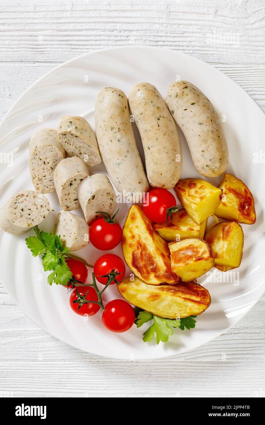 Weisswurst, bavarian white sausage of minced veal, pork back bacon, spices and parsley on white plate with roast potatoes, fresh tomatoes, vertical vi Stock Photo