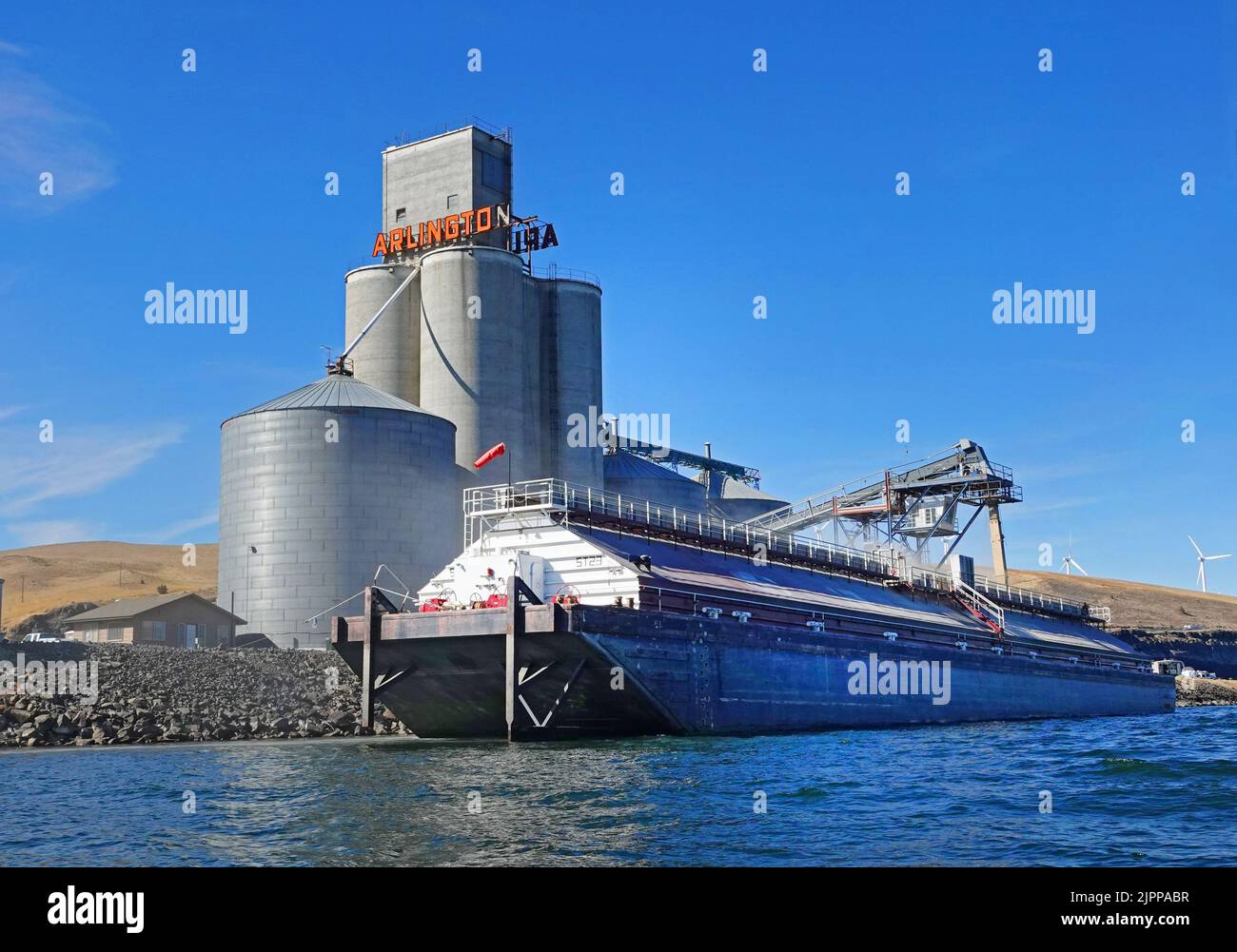 A barge on the Columbia River in the tiny port of Arlington, Oregon takes on a load of freshly cut wheat from a massive, shoreside silo. Stock Photo