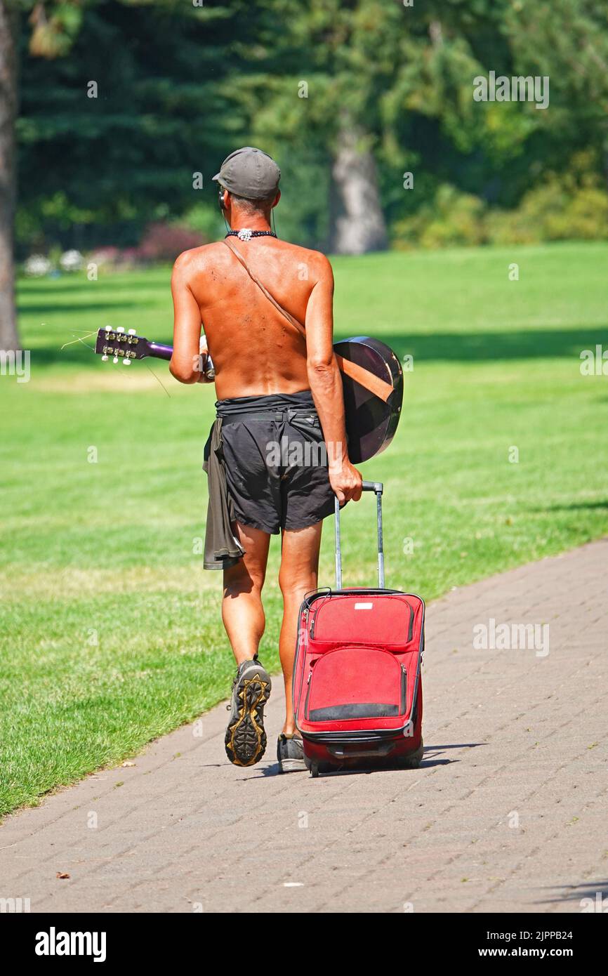 A man wearinfg only shorts and a guitar strolls along the Deschutes River in Bend, Oregon, during the July/2022 heat wave that brought 100 degree plus temperatures to the Pacific Northwest. Stock Photo