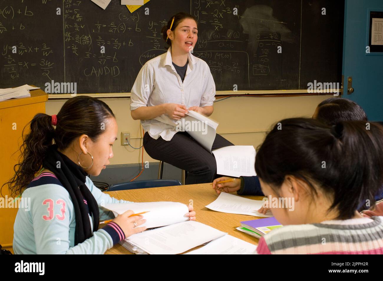 Education High School female teacher talking to class, pencil behind her ear, female students following subject with papers on table Stock Photo