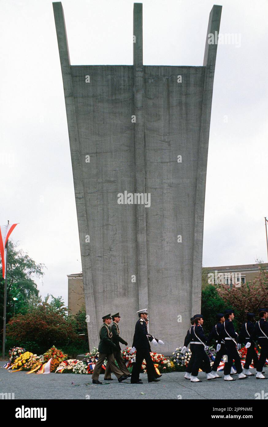 A French honor guard, followed by German policemen, marches past the Berlin Airlift Memorial during a ceremony honoring those who lost their lives during the Berlin Airlift of 1948-1949. The arc of the memorial points to the West and the prongs at the top represent the three air routes to and from West Germany, circa 1984 Stock Photo