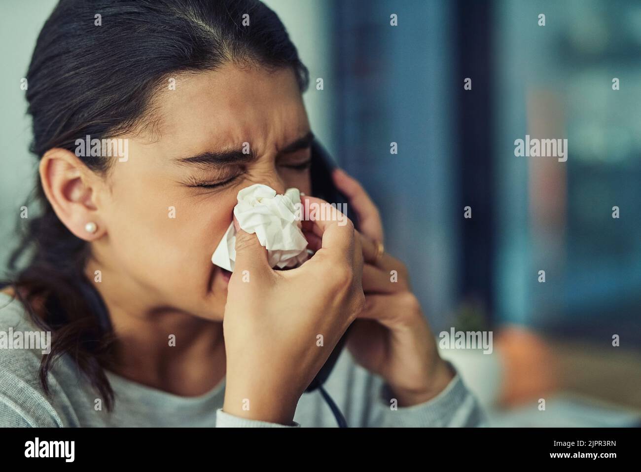 Suffering with uncontrollable sneezes. a young businesswoman blowing her nose while speaking on a phone in an office. Stock Photo