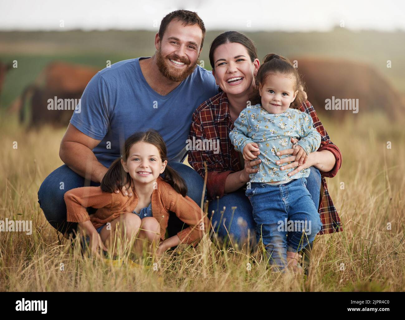 Portrait of happy family on a countryside farm field with cows in the background. Farmer parents bonding with kids on a sustainable agriculture cattle Stock Photo