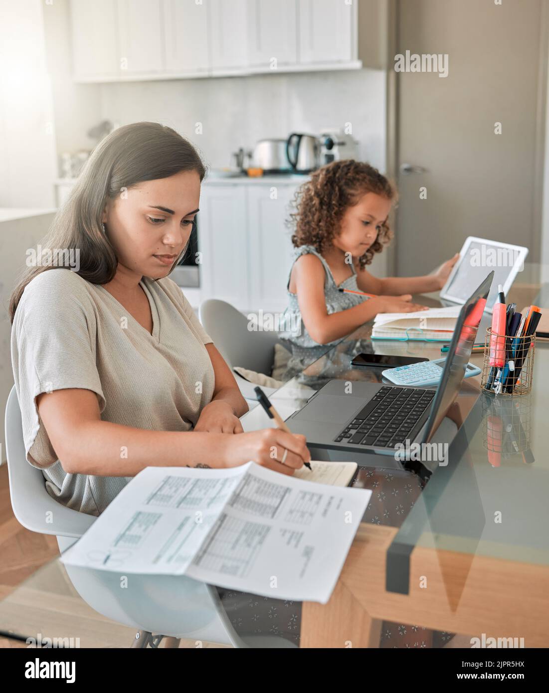 Mother and daughter being productive with remote work and homework, multitasking at a kitchen table at home. Parent and child serious while paying Stock Photo
