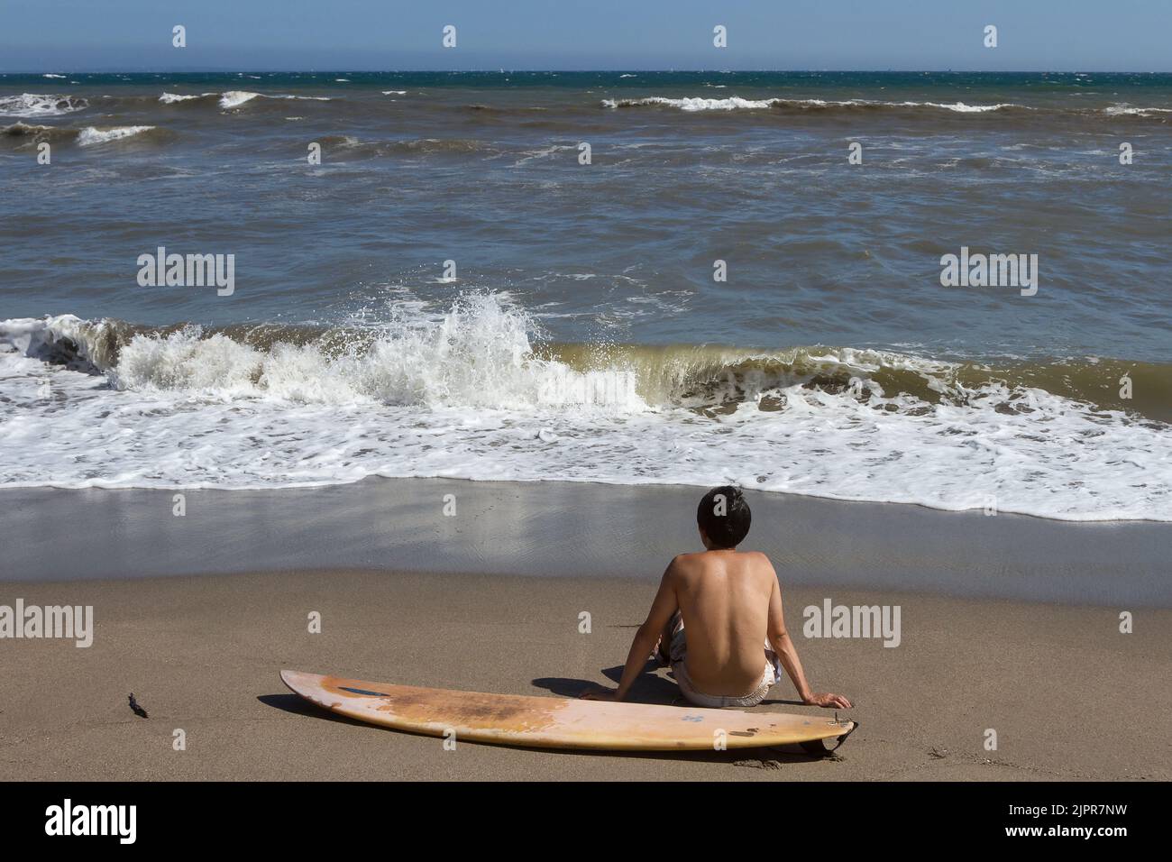 A surfer sit by his surfboard on the beach watching the waves. Enoshima, Kanagawa, Japan. Stock Photo